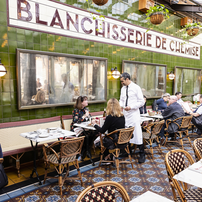 Vintage-style restaurant with green tiled walls, formal seating, and a waiter serving guests. "BLANCHISSEURIE DE CHUMISE" sign.