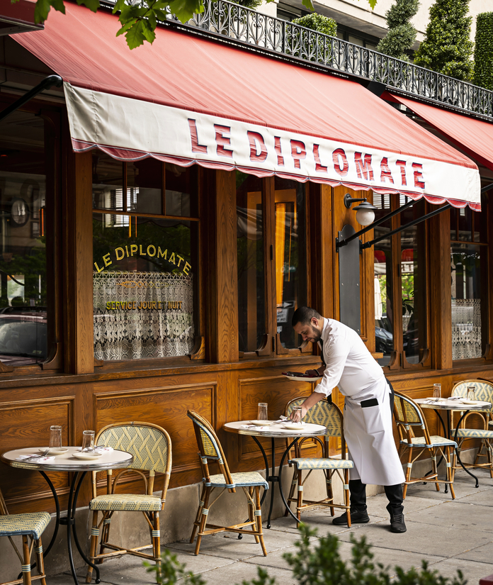 Waiter in white uniform setting outdoor table at Le Diplomate restaurant with red awning, wooden facade, and greenery.