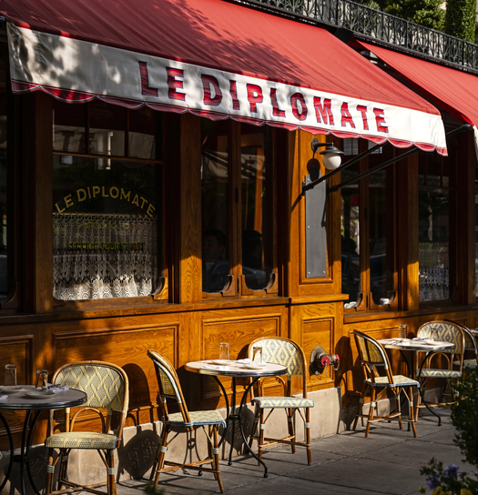 A bright outdoor cafe scene with red awning, "Le Diplomate" signage, wooden tables, chairs, and greenery.
