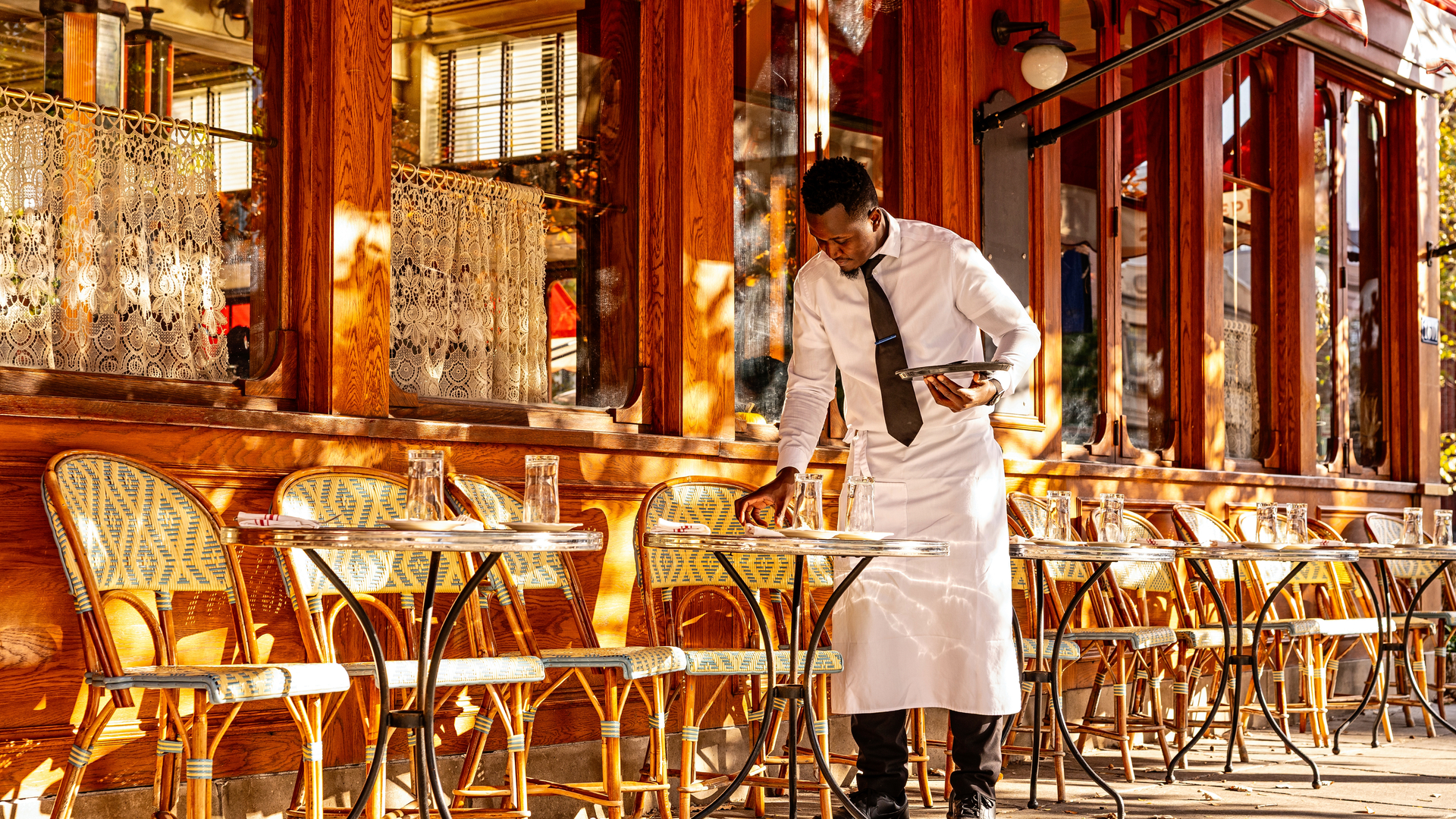 Waiter in white uniform and black tie adjusts tablecloth at outdoor cafe with wooden chairs and阳光。