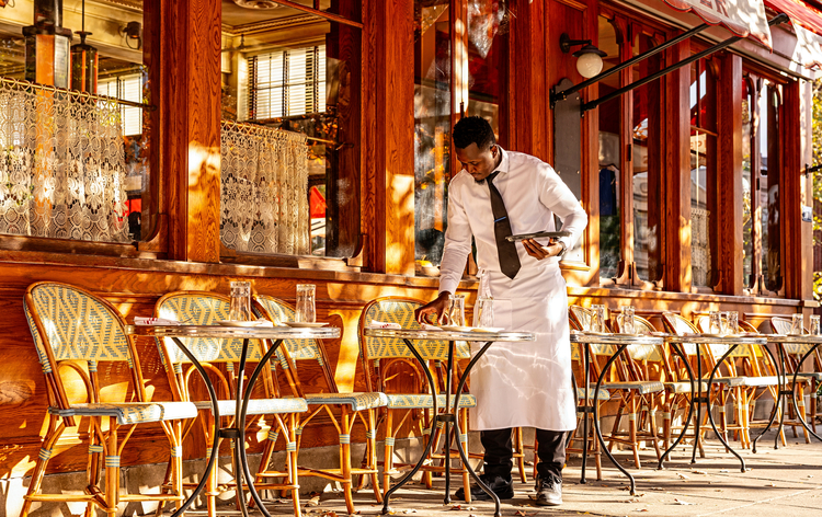 A server in a white apron stands at a outdoor cafe table, sunlit by morning light, with wooden chairs and windows in the background.