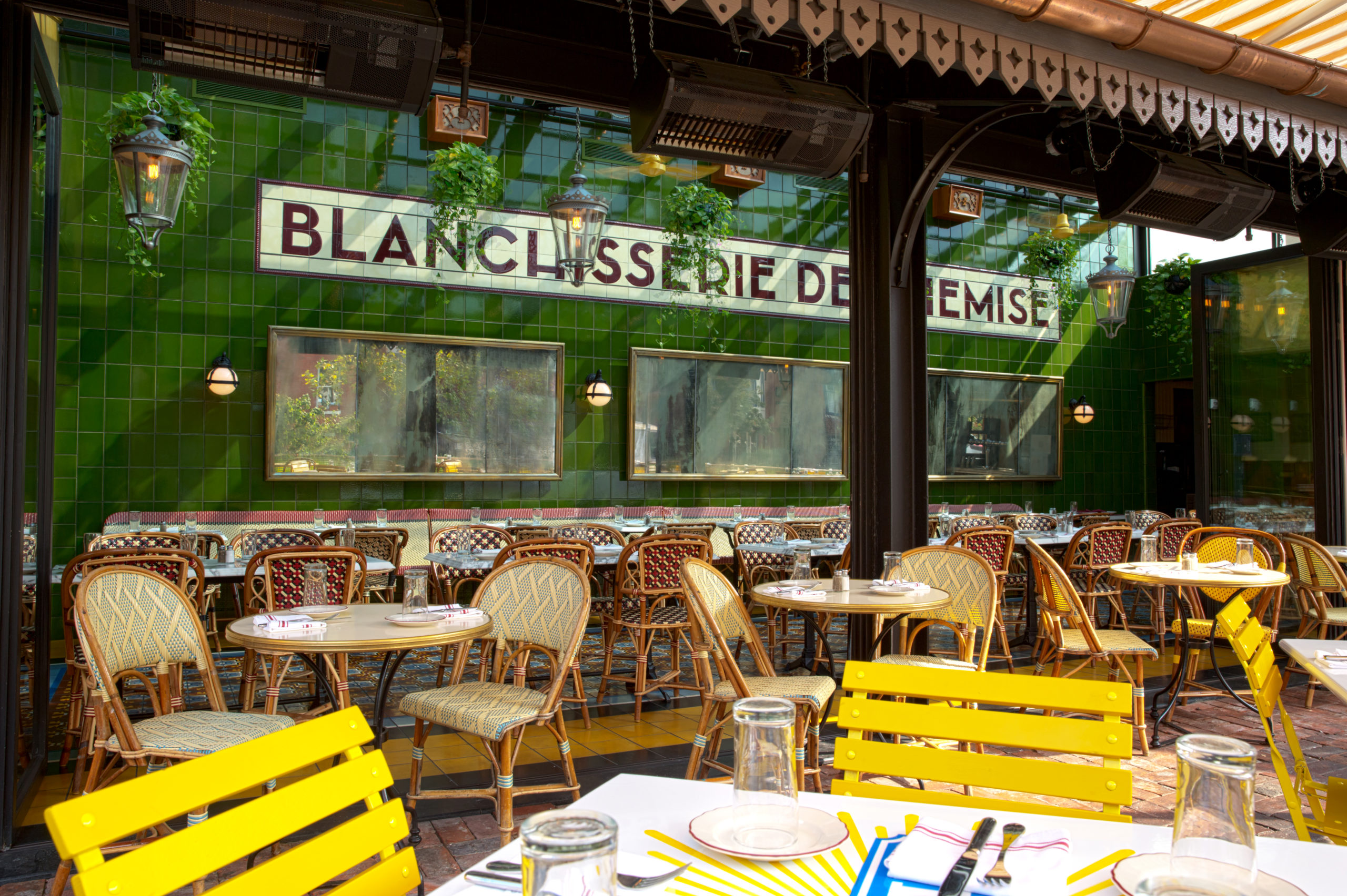 View of outdoor seating - green tile walls, tile and brick flooring, red/white, blue/tan, yellow chairs. Greenery hanging.